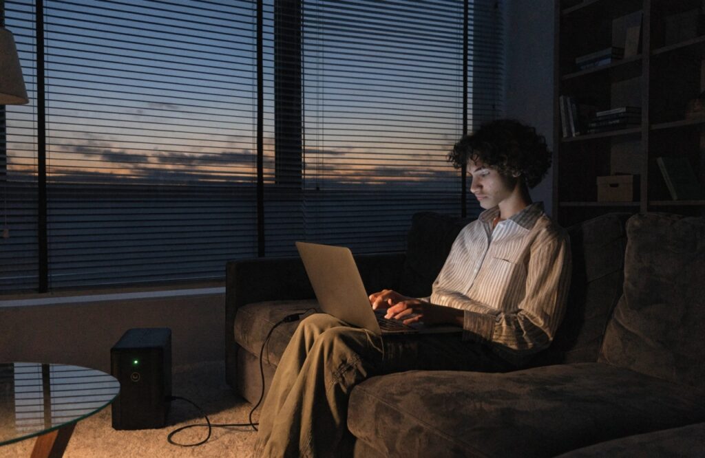 A person working comfortably on a laptop in a dimly lit room, with the laptop's power adapter plugged into a UPS on the floor